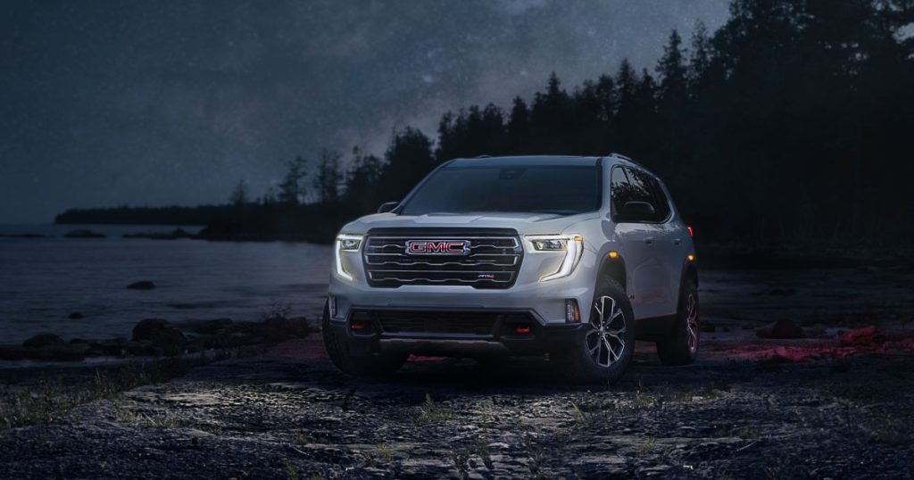 A GMC Acadia parked by a lake off-road at night with the headlights on.