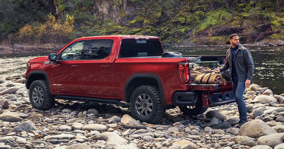 A red 2026 GMC Sierra 1500 pickup truck parked off-road by a river with the owner packing camping gear in the truck's bed.